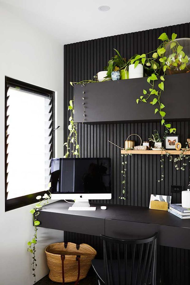 A compact, stylish home office with a black built-in desk against a dramatic vertical slatted feature wall. The workspace includes a desktop computer, keyboard, books, framed photographs, open shelving, and abundant trailing indoor plants. A louvred window provides bright natural light, while a black stool and woven basket add functional furnishings.
