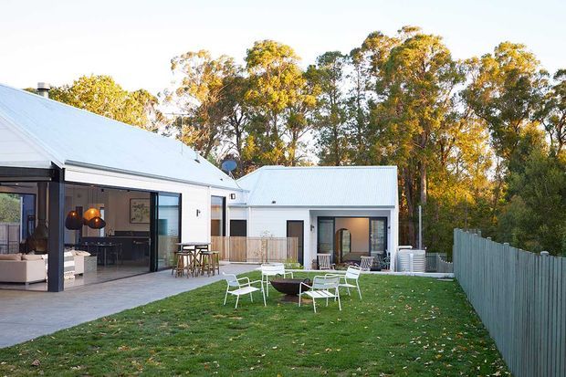 A contemporary single-storey residential property with white-painted walls, light grey corrugated metal pitched roofs, and extensive glazed openings. The image looks across a paved patio and lawn toward the main house and a smaller detached or connected wing, with outdoor dining and lounge furniture in the foreground. Mature trees and a simple vertical timber fence surround the garden.