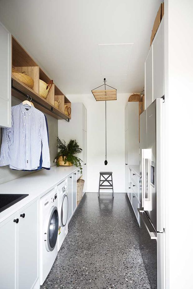 A bright, narrow laundry room with white cabinetry and benchtops, integrated front-loading washer and dryer, open timber shelving with woven baskets, hanging clothes, and a dark terrazzo-style floor. A simple timber pendant light and indoor plant add warmth to the functional space.