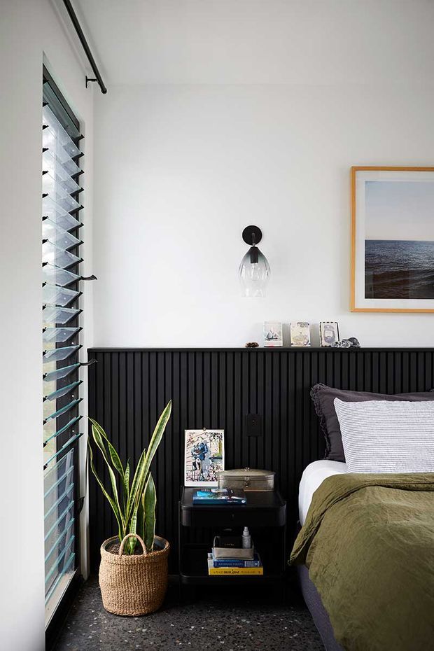 A compact, stylish bedroom corner with a bed dressed in muted olive-green and grey bedding, a black fluted half-height wall, and a small black bedside table with books and decor. A tall indoor plant sits in a woven basket beside full-height louvred glazing, while a clear pendant light and framed ocean photograph add visual interest. The room has a calm, contemporary feel with strong black-and-white contrast and natural accents.