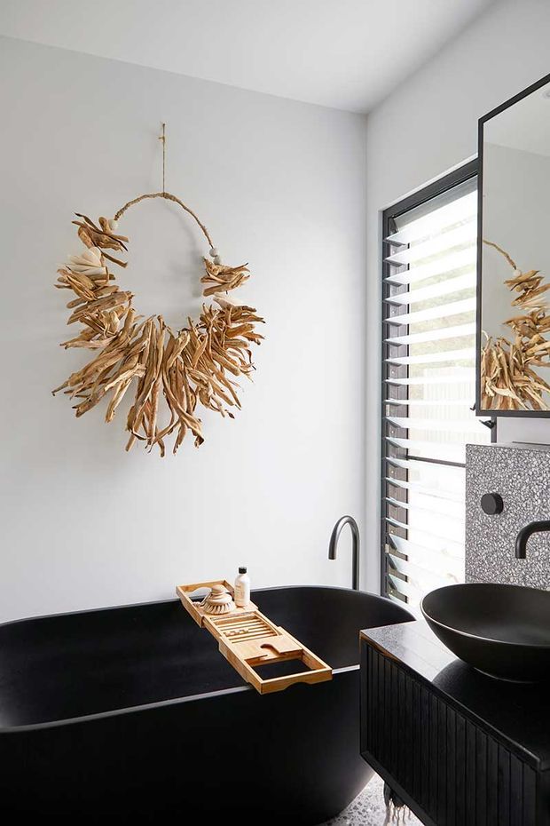 A refined contemporary bathroom featuring a deep matte-black freestanding bathtub, matching black vessel basin and vanity, a dark textured wall panel, and a slim black-framed louvred window. White walls provide contrast, while a natural dried botanical wreath and timber bath tray add organic warmth.
