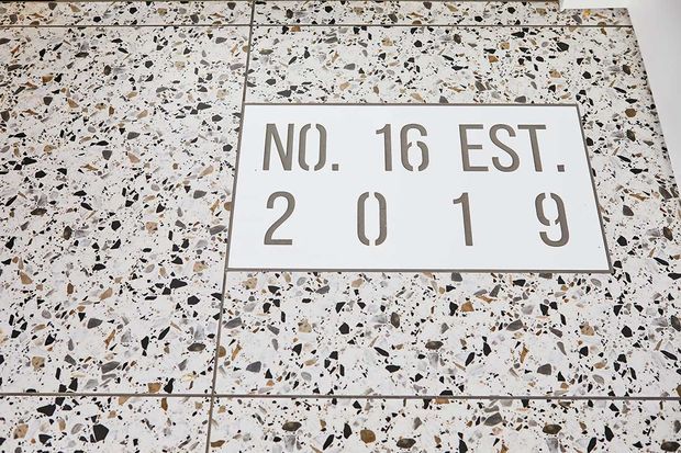 Close-up of a terrazzo-clad wall or facade featuring a recessed rectangular sign reading “NO. 16 EST. 2019.” The terrazzo has a light base with black, grey, and warm brown aggregate chips, arranged in large square panels with visible grout lines.