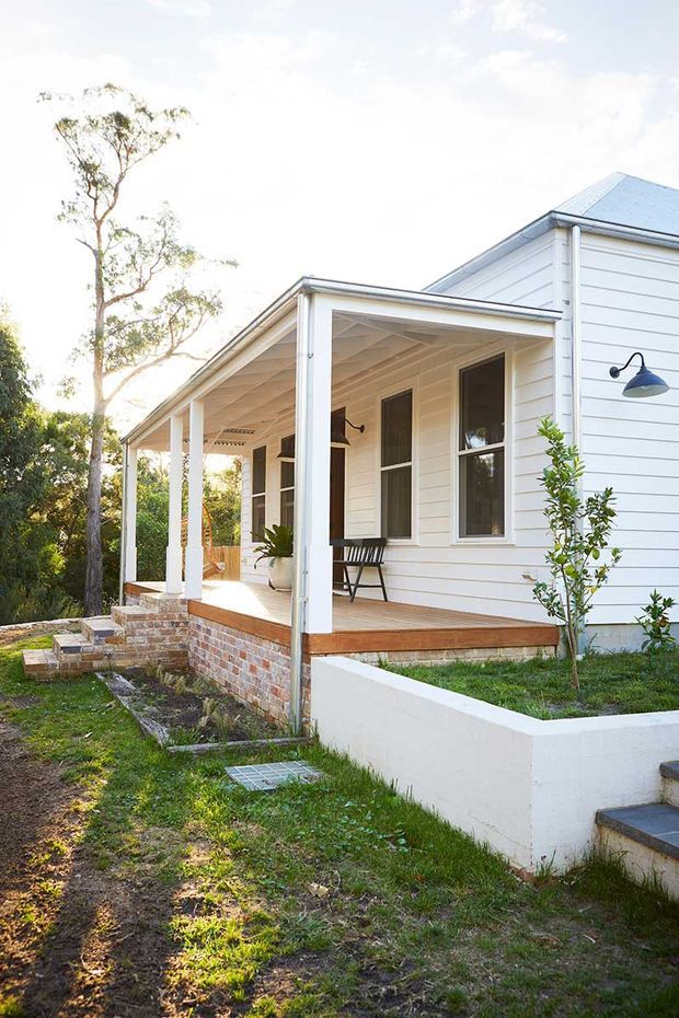 Exterior view of a bright white weatherboard house with a covered front verandah, timber decking, brick base, pitched metal roof, and simple dark wall-mounted lights. The landscaped front garden includes low rendered retaining walls, steps, lawn, and established trees.