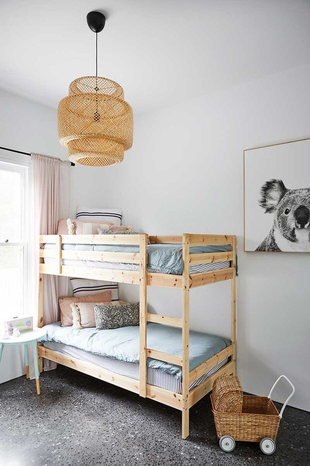 Bright children's bedroom featuring a natural timber bunk bed with pale blue bedding, blush and patterned cushions, a woven rattan pendant light, soft pink curtains, koala artwork, a small side table, and a woven toy pram. The room has white walls and a dark speckled floor.
