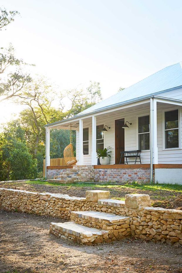 Sunlit exterior view of a single-storey white weatherboard house with a broad covered front veranda, pale metal pitched roof, brick and stone foundations, timber-framed windows, and a hanging wicker chair. Rustic stone retaining walls and steps lead up from the gravel foreground, with mature trees surrounding the property.
