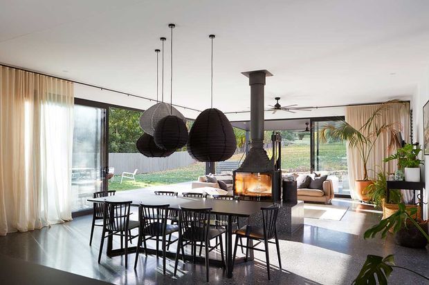 Open-plan contemporary living and dining area with full-height glazing, sheer curtains, a large dark dining table, sculptural pendant lights, and a central suspended wood-burning fireplace. The room opens to a landscaped outdoor setting and features abundant indoor plants and neutral upholstered seating.