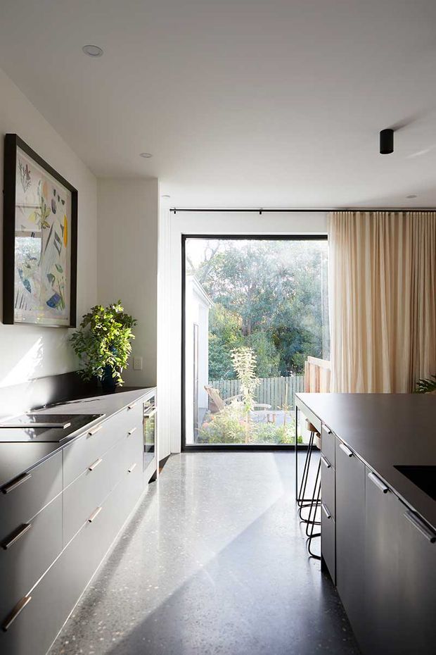 Bright, contemporary kitchen with parallel runs of sleek white cabinetry, dark benchtops, and a long central island with bar stools. A full-height glazed door opens to a leafy outdoor area, framed by beige curtains. The room features polished grey flooring, white walls and ceiling, black window and lighting details, integrated appliances, and a large framed artwork.
