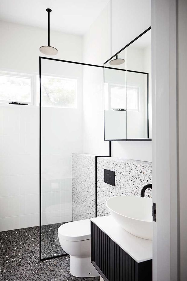 Bright contemporary bathroom with a frameless glass shower enclosure outlined by slim black framing, a wall-mounted mirror cabinet, white vessel basin, toilet, black fluted vanity cabinetry, and terrazzo-style flooring and wall surfaces. The room is predominantly white with strong black accents and abundant natural light.
