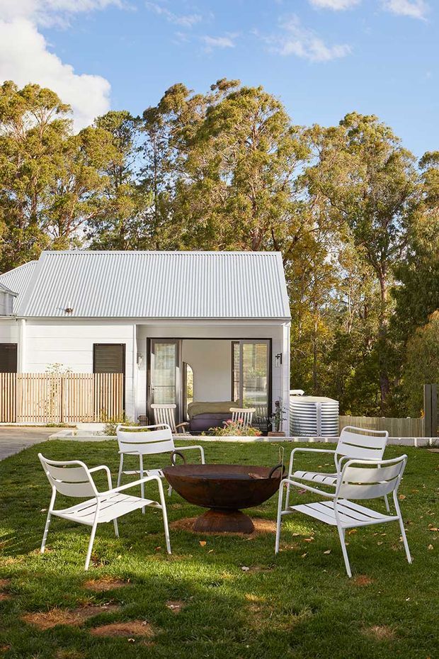 A small white cottage-style house with a corrugated metal gable roof sits within a leafy garden. The foreground features a circular rusted metal fire bowl surrounded by white outdoor lounge chairs on a lawn. Large glazed openings, a small covered porch, timber screening, and a rainwater tank are visible against a backdrop of mature trees.