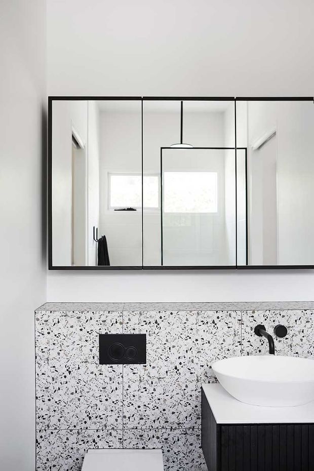 A bright contemporary bathroom featuring a wall-mounted black-framed mirrored cabinet, white wall tiles, a terrazzo-look tiled vanity wall, a white vessel basin on dark ribbed cabinetry, and black tapware and flush plate. A toilet is partially visible at the bottom of the frame.