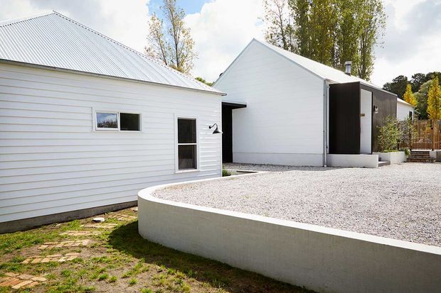 Exterior view of a minimalist cluster of low-rise buildings with white horizontal cladding, simple pitched metal roofs, and a dark vertical-clad entrance volume. A curved raised concrete planter or terrace filled with pale gravel occupies the foreground, with mature trees and timber fencing in the background.