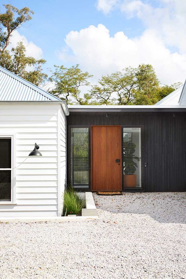 Exterior view of a low-rise residential entrance combining a white horizontal weatherboard wing with a dark vertically ribbed facade. A warm timber front door is flanked by narrow glazed sidelights, with pale gravel landscaping, a small planted bed, corrugated metal roof forms, and mature trees beyond.