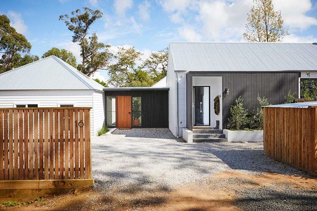 Single-storey residential property viewed from the front, featuring a contemporary low-profile composition with two pitched corrugated metal roof forms, dark vertical cladding, light metal roof sheeting, timber entry doors, timber slat fencing, gravel paving and simple planted beds. Mature trees surround the house, and the partially enclosed entry courtyard creates a private arrival space.