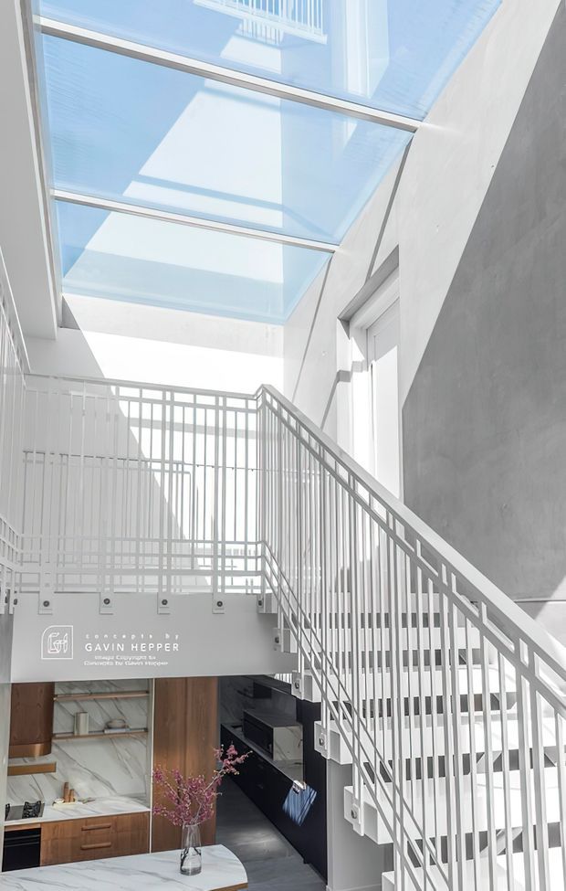 Bright contemporary multi-level interior with a white metal stair balustrade, open stairwell, large overhead skylight, and a double-height void. A compact kitchen with dark cabinetry, timber joinery, and a marble-look surface is visible below, while white walls and abundant daylight create an airy architectural composition.