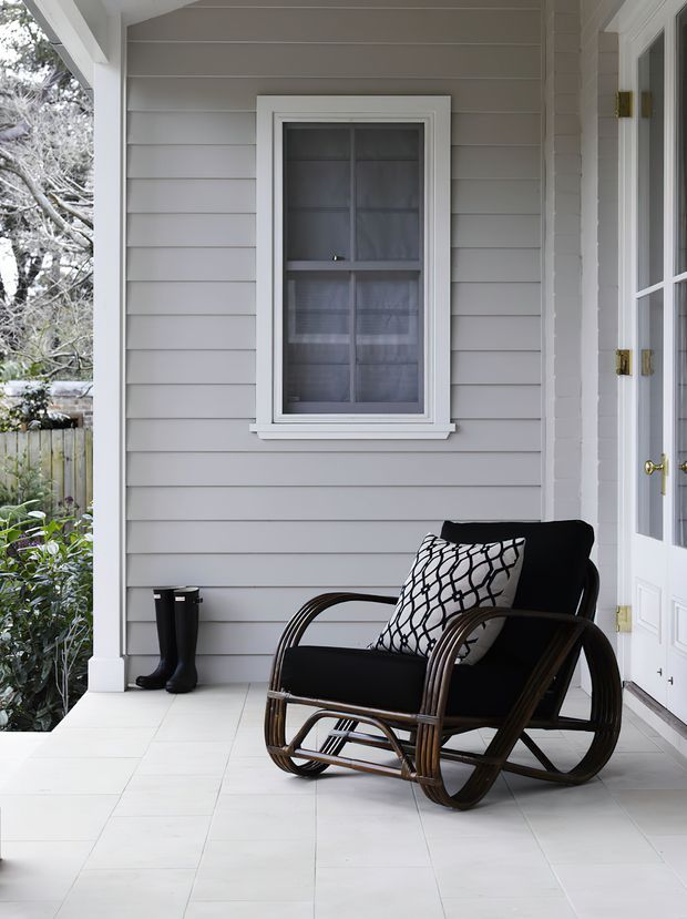 Covered residential front porch with pale grey horizontal weatherboard walls, white trim, a divided-light window, and glazed double doors. A dark rattan rocking chair with patterned cushion sits on a light tiled floor beside a pair of black boots, creating a calm, traditional entrance setting.