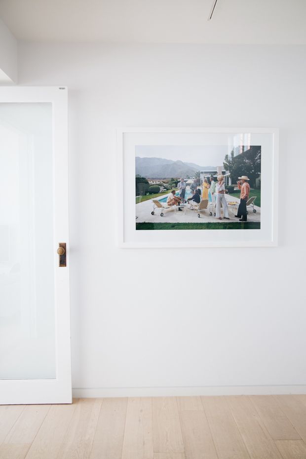 Minimal, bright interior detail showing a white wall, pale timber floorboards, and a white-framed glazed door with a brass-toned handle. A large white-framed artwork hangs on the wall, while the mostly empty composition emphasizes clean lines and abundant natural light.