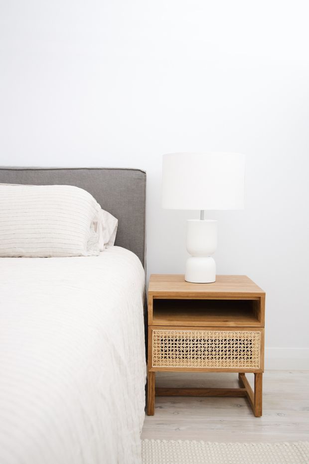 Bright, minimalist bedroom detail showing the edge of a neatly made bed with white striped bedding and a grey upholstered headboard. A natural timber bedside table with woven cane-front storage stands beside it, topped with a simple white ceramic lamp. Pale timber flooring, white walls, and a light textured rug create an airy, calm interior.