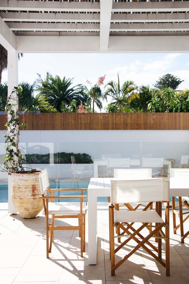 Bright outdoor dining terrace beneath a white pergola, furnished with a white dining table and wooden director-style chairs. Full-height glazing reflects the garden and reveals a swimming pool beyond, while a timber privacy fence, tropical planting, and a large weathered ceramic planter define the setting.
