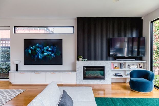 Bright contemporary living room featuring two wall-mounted televisions, a long white built-in media console, a black vertical-slat feature wall, a stone-clad fireplace, open shelving, timber flooring, and a deep blue accent armchair. Large windows and high-level glazing bring in natural light, while a teal rug adds colour.