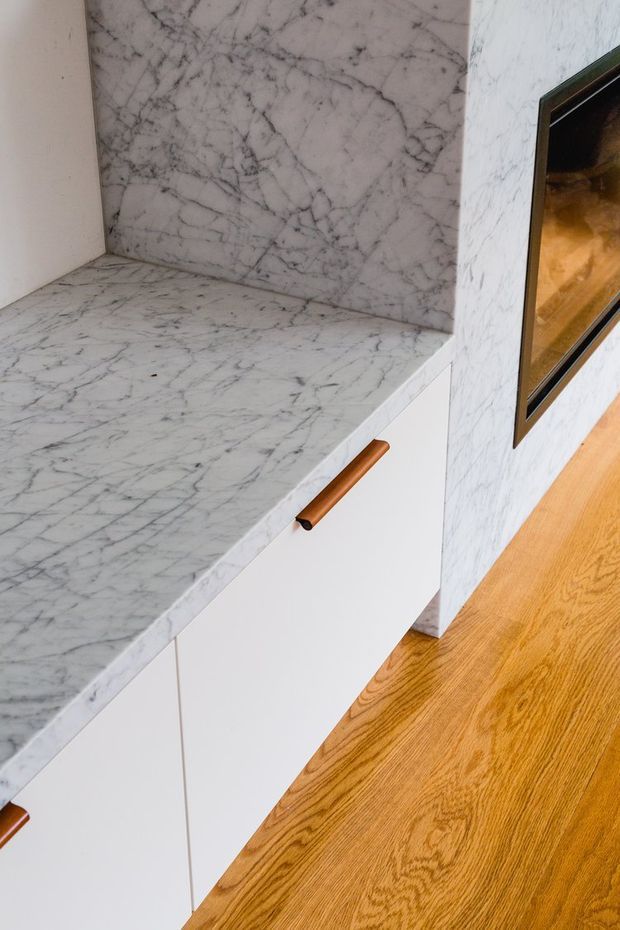 Close-up interior view of custom white cabinetry with a white-grey veined marble benchtop and matching marble backsplash. A warm timber pull handle, integrated dark-framed appliance or fireplace opening, and honey-toned timber flooring are visible.