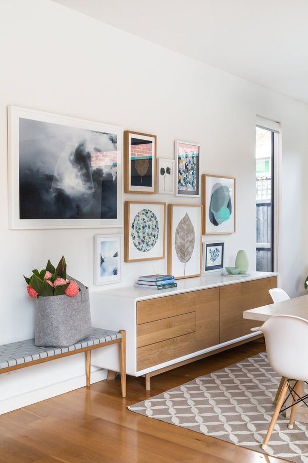 Bright, carefully styled mid-century-inspired living and dining area with white walls, warm timber flooring, a low wooden sideboard, framed botanical and abstract artwork, a bench seat, and a patterned rug. A dining table and molded white chairs are partially visible on the right, while a tall window brings in natural light.