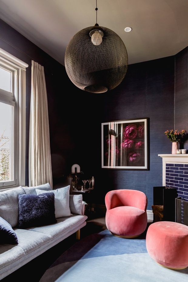 A richly styled living room with dark navy-black wall panelling, a pale ceiling, tall window with light curtains, and a large woven spherical pendant light. A pale sectional sofa, plush coral-pink rounded lounge chairs, a blue-toned area rug, framed artwork, drinks table, and a fireplace with dark blue tile surround create a dramatic, eclectic interior.