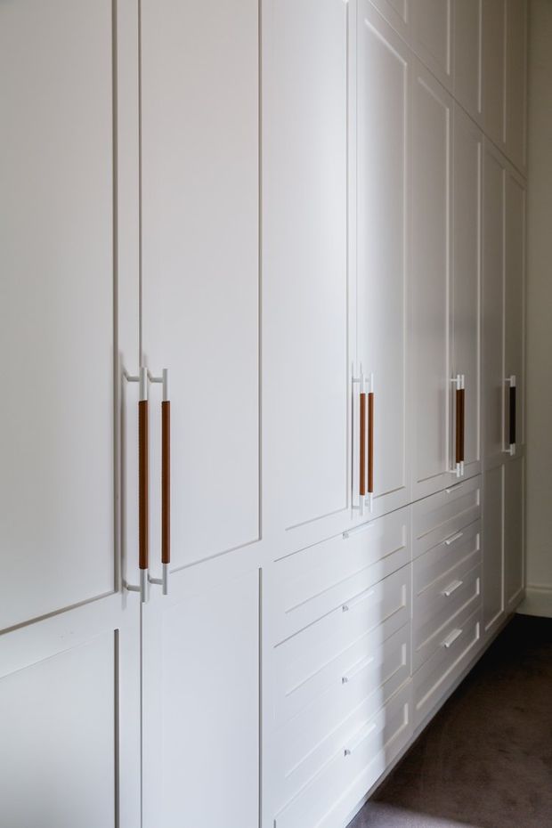A long wall of tall, floor-to-ceiling white built-in cabinetry with recessed panel doors and integrated drawers. Slim paired handles combine warm timber-toned grips with light metal fittings. The cabinetry lines a narrow, softly lit interior with dark carpet flooring, creating a highly ordered storage wall.