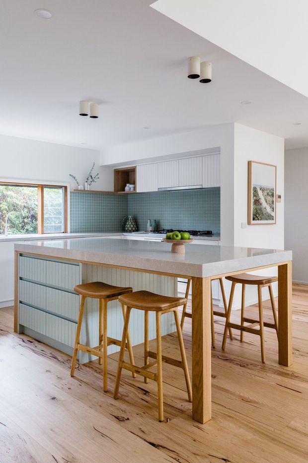 Bright contemporary kitchen with a large island and integrated dining-height extension, pale timber flooring, muted blue-green tiled splashback, light blue-grey vertical-panel cabinetry, and warm timber stools and table legs. White ceilings and walls, timber-framed glazing, and simple cylindrical ceiling lights create an airy, refined space.