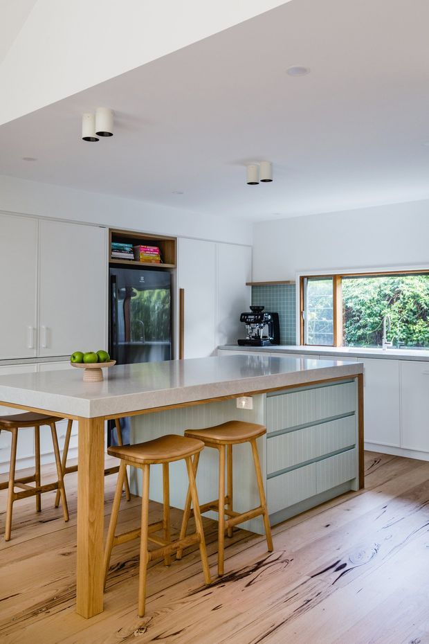 Bright contemporary kitchen featuring a large pale stone island with a timber breakfast-bar extension, integrated white cabinetry, a dark integrated refrigerator, ribbed muted green-grey island cabinetry, timber stools, and a dark teal square-tile splashback. Natural light enters through a timber-framed window overlooking greenery. The room has pale timber flooring and simple white recessed and cylindrical ceiling lighting.