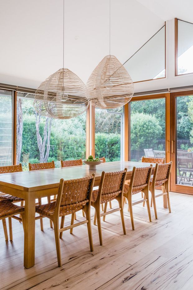 Bright dining room with a long timber dining table and woven leather dining chairs, surrounded by extensive timber-framed glazing overlooking greenery. Three large woven pendant lights hang above the table, while a high angled ceiling and large upper window add an airy feel. Pale timber flooring and warm wood tones create a natural, inviting interior.
