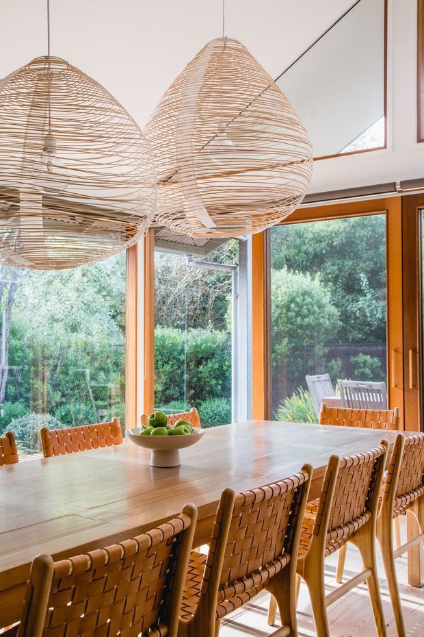 A bright dining room centered on a large rectangular timber dining table surrounded by woven leather or rattan dining chairs. Three oversized woven pendant lights hang above the table. Floor-to-ceiling timber-framed glazing opens views to a lush garden, creating an airy connection to the outdoors. The room has warm natural materials, light timber flooring, and a relaxed contemporary feel.