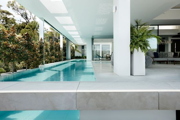 A bright contemporary indoor-outdoor living space featuring a long narrow turquoise swimming pool alongside full-height glazing and a covered terrace. White structural columns, a white ceiling with rectangular skylights, pale stone or concrete flooring, lush planting, and a partially visible lounge area create a clean, resort-like atmosphere.