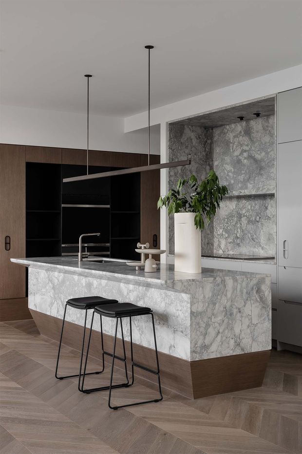A sophisticated contemporary kitchen centered on a large marble-clad island with an integrated sink and two slim black bar stools. Dark timber cabinetry with black open shelving lines the rear wall, while a large veined stone feature panel and tall light cabinetry add contrast. Minimal linear pendant lights hang above the island, and the herringbone timber floor creates a warm foundation.