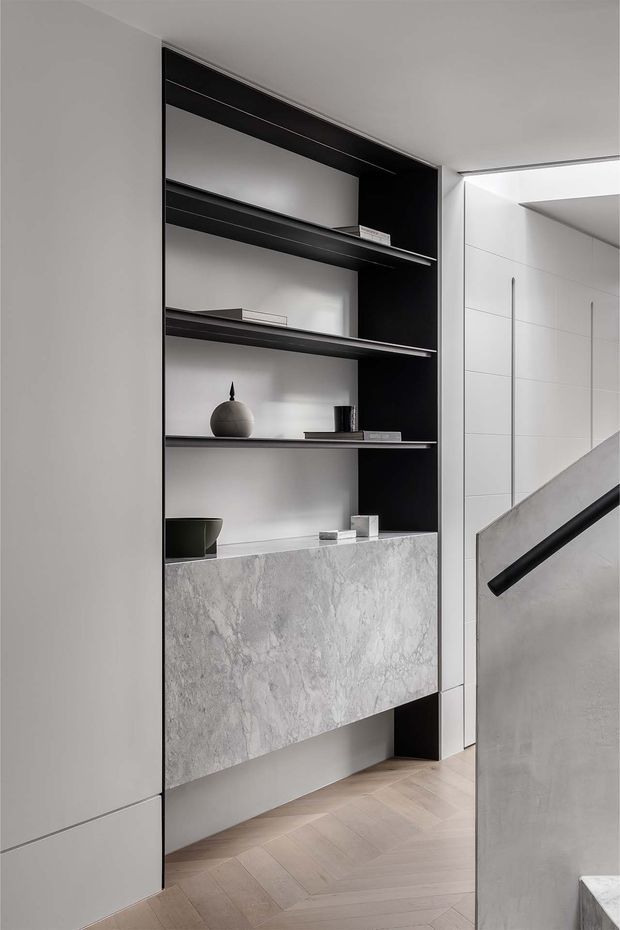 A refined contemporary interior detail showing a built-in black and white shelving unit with open display shelves and a large grey stone-faced drawer or cabinet. Pale timber herringbone flooring runs through the space, while a concrete stair balustrade with a black handrail is visible at right. The palette is monochromatic and minimalist, with carefully styled neutral objects on the shelves.