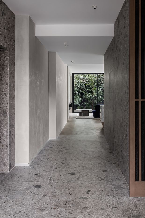 A long, refined interior hallway with large-format grey terrazzo or natural stone flooring, textured grey wall finishes, white ceilings, and dark timber-framed screening at the right. The corridor leads toward a glazed living area with leafy greenery visible outside. The space has minimalist detailing, recessed lighting, and a restrained neutral palette.