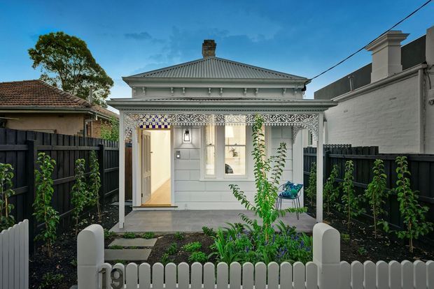 Front exterior view of a compact heritage-style cottage with a pale painted facade, hipped corrugated metal roof, ornate decorative verandah detailing, timber-framed windows and entry door. A small landscaped garden sits behind a white picket fence, with dark boundary fencing on both sides.