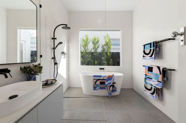 Bright contemporary bathroom with a freestanding white bathtub positioned beneath a large window, a black wall-mounted shower, double vanity with vessel basins, and open black towel rails displaying colourful patterned towels. Large-format grey floor tiles, pale walls, and a frameless mirror create a clean, spacious feel.