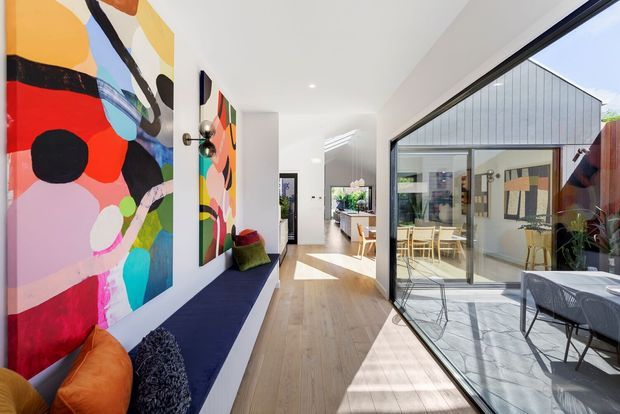 Bright contemporary interior hallway or open-plan circulation space with a long built-in navy bench, colourful abstract artwork, timber flooring, white walls and ceiling, and extensive black-framed floor-to-ceiling glazing. The glazing opens visually onto a sunlit courtyard or patio with outdoor dining furniture and a pitched-roof building beyond.