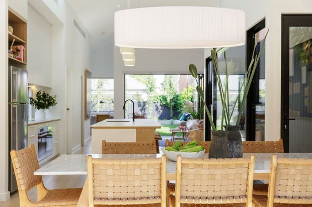 Bright open-plan kitchen and dining area with a white stone island and integrated sink, stainless-steel appliances, pale cabinetry, a large white pendant light, and a timber dining table with woven leather or cane-style chairs. Full-height glazing and a glazed door open toward a leafy courtyard or garden, while tall indoor plants add a natural accent.