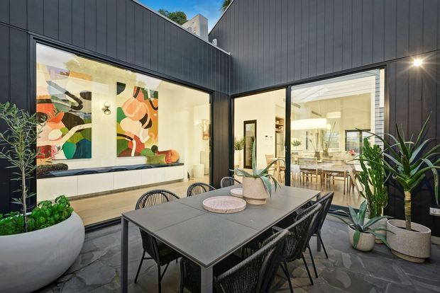 A contemporary inner courtyard dining area enclosed by dark vertical-panel cladding. A large outdoor dining table with woven black chairs sits among potted plants on irregular stone paving, with expansive glazing opening views into a bright, art-filled living area and dining space beyond.