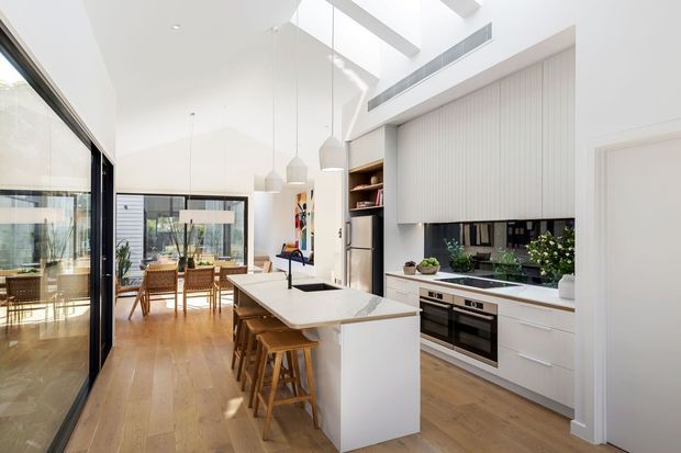 Bright, contemporary open-plan kitchen with a large white island and integrated sink, timber bar stools, white cabinetry, stainless-steel appliances, and a vertically fluted grey feature wall. Pendant lights, skylights, and clerestory glazing create an airy connection to the adjacent dining area, which is visible beyond the kitchen.