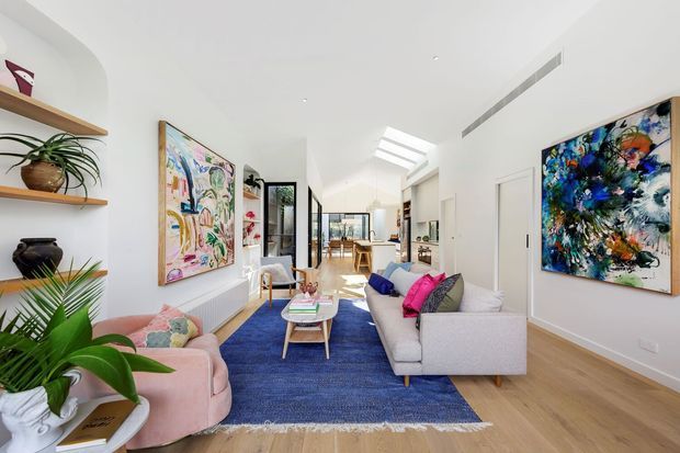 Bright contemporary open-plan living room with white walls, light timber flooring, a large skylight, colourful abstract artwork, built-in shelving with plants, and a pale sectional sofa arranged around a coffee table on a blue rug. The space extends toward a dining and kitchen area at the rear.