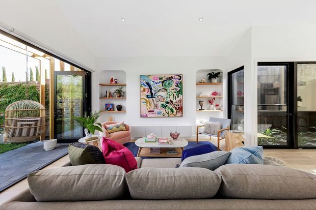 Bright contemporary living room viewed from behind a large sectional sofa, with floor-to-ceiling black-framed glazing opening to an outdoor deck, built-in white shelving niches, colourful artwork, a coffee table, and layered cushions. The room has a light, airy palette with vivid pink and blue accents and strong indoor-outdoor connection.
