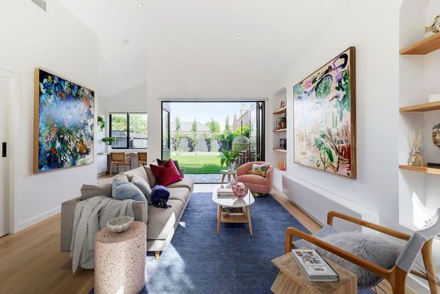 Bright contemporary living room with white walls, timber flooring, a large blue area rug, sectional sofa, sculptural coffee tables, timber-framed armchairs, open display shelving, and colourful abstract artwork. Full-height glazing opens the room to a landscaped backyard, creating an airy connection to the outdoors.