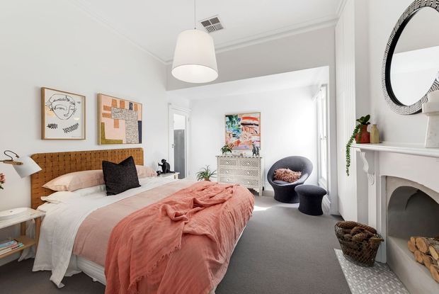 Bright, inviting bedroom with a textured tan upholstered headboard, layered white and blush-pink bedding, framed artwork, a compact dresser, sculptural lounge chair, and a decorative fireplace with a large round mirror. The room has white walls and ceiling detailing, grey carpet, and a soft contemporary eclectic styling.