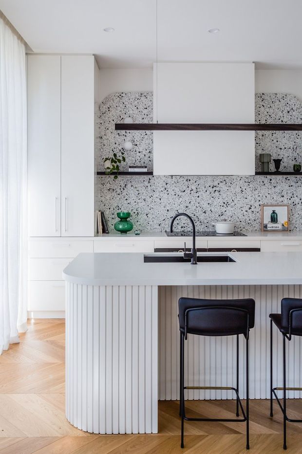 Bright contemporary kitchen with a large white island, integrated sink and dark gooseneck tap, fluted vertical island cladding, white cabinetry, and a terrazzo-style speckled backsplash. Dark timber open shelving and black bar stools add contrast, while full-height sheer curtains and warm timber parquet flooring create an airy, refined interior.
