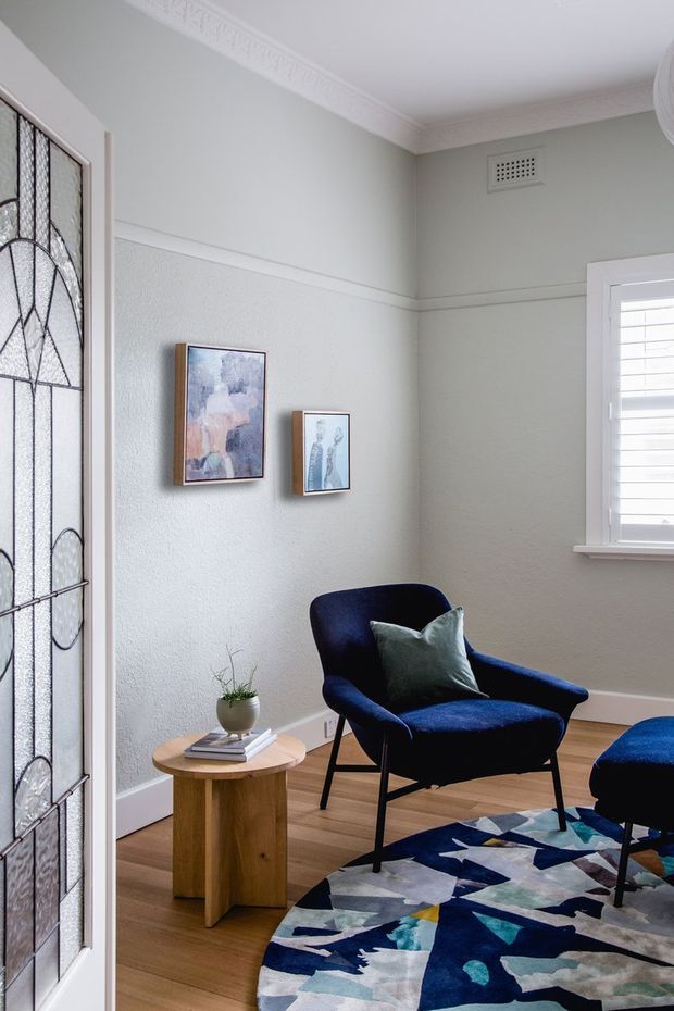 A refined sitting area in a period-style home, featuring pale grey-green walls with decorative picture rails and ornate ceiling cornice, timber flooring, a stained-glass entry door, plantation-shuttered window, navy upholstered armchair and ottoman, a small timber side table, framed artwork, and a bold geometric area rug.