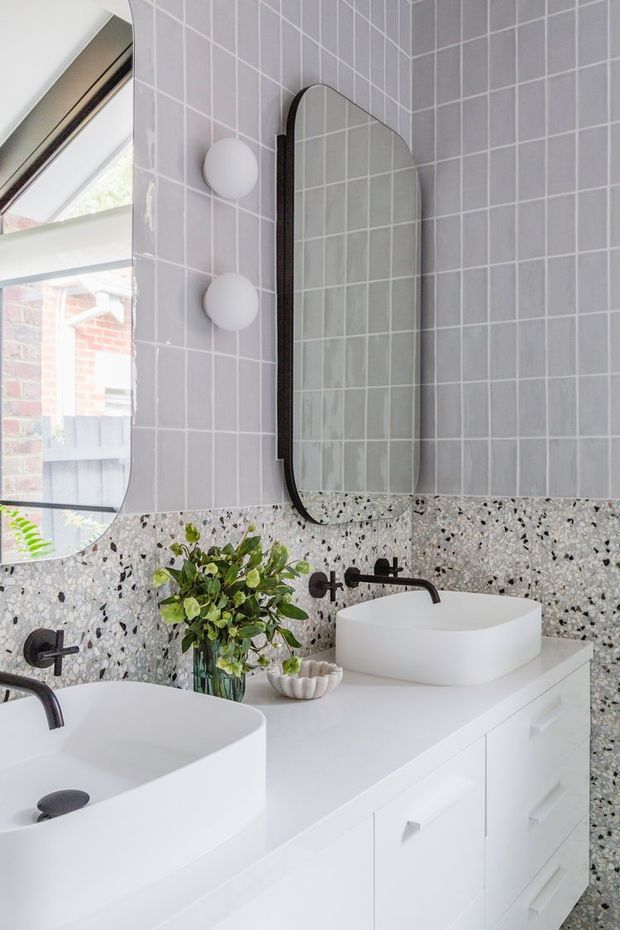Bright contemporary bathroom vanity featuring a double white vessel sink, white built-in cabinetry, dark wall-mounted tapware, pale square wall tiles, a terrazzo-style feature backsplash, and rounded-edge mirrors with globe wall lights.