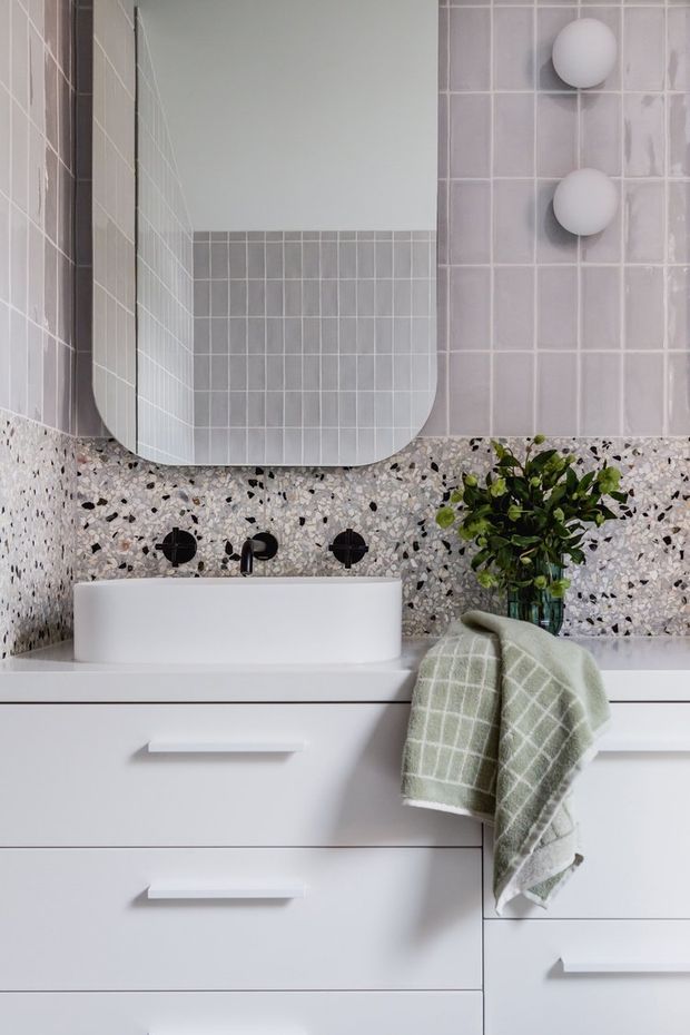 Close-up view of a contemporary bathroom vanity with a white vessel basin, matte black wall-mounted tapware, white cabinetry, pale grey square wall tiles, terrazzo splashback and benchtop surfaces, a rounded rectangular mirror, and a green towel draped over the drawers. A small plant adds natural contrast.