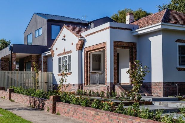 Exterior view of a renovated heritage-style brick and rendered residence, combining an older tiled-roof frontage with a contemporary rear extension. The facade features decorative brick detailing, white rendered walls, contrasting dark modern forms, steel-framed windows and a landscaped front garden behind low brick walls.